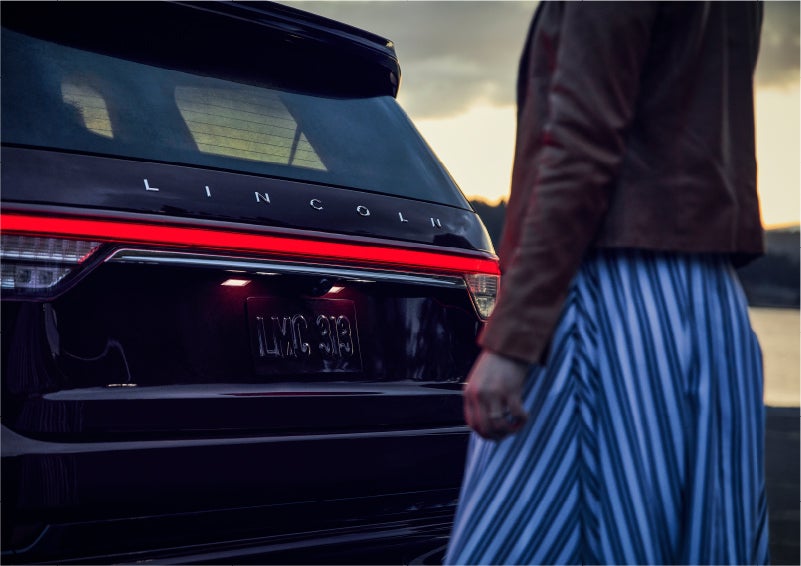 A person is shown near the rear of a 2023 Lincoln Aviator® SUV as the Lincoln Embrace illuminates the rear lights | Boulevard Lincoln in Georgetown DE