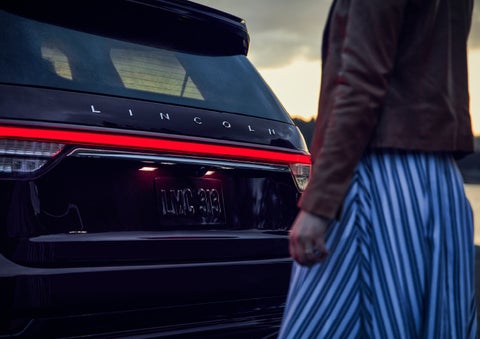 A person is shown near the rear of a 2024 Lincoln Aviator® SUV as the Lincoln Embrace illuminates the rear lights | Boulevard Lincoln in Georgetown DE